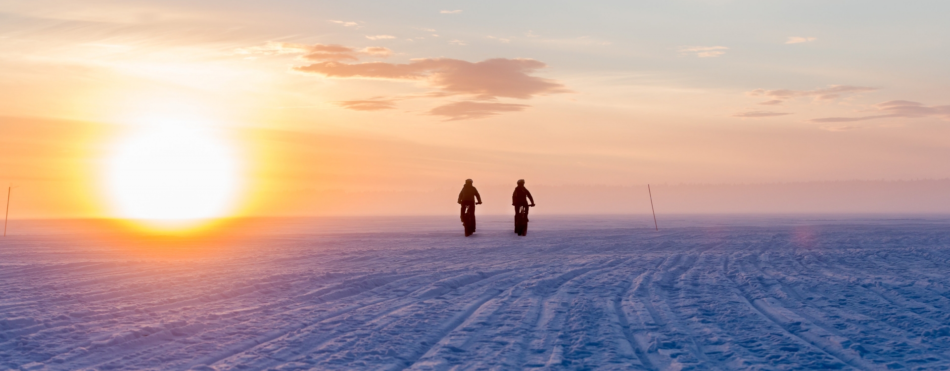 Two silhouetted figures on bikes riding across a snowy expanse toward a large glowing sun at sunset, warm orange and purple tones across a vast winter landscape