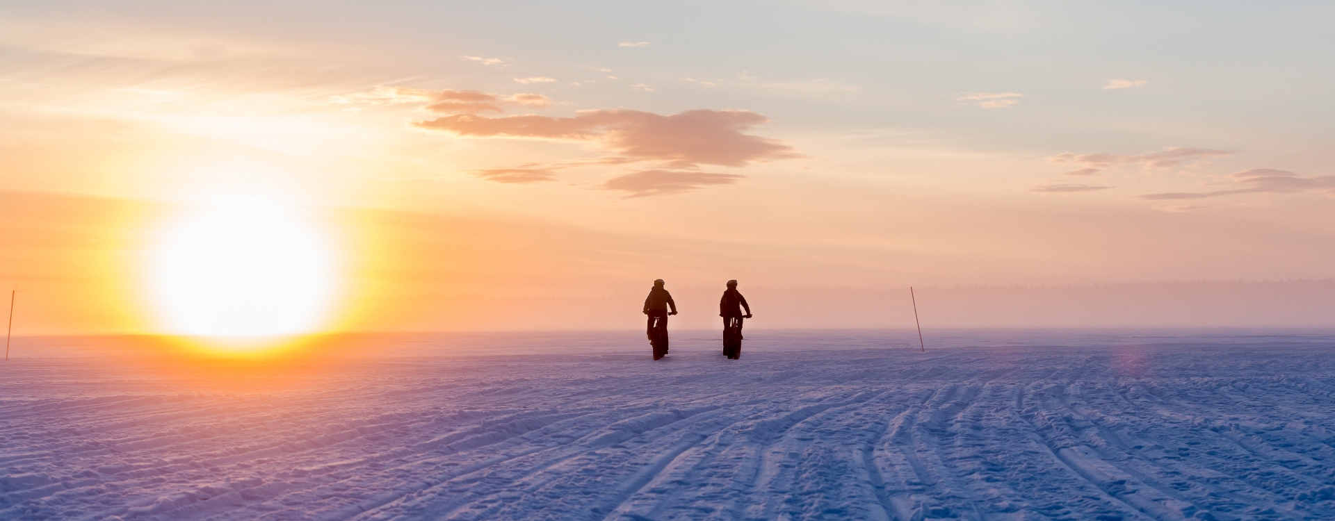 Two silhouetted figures on bikes on a snowfield near a bright setting sun, orange and lavender sky, peaceful atmosphere